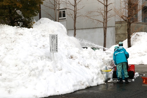 人の背丈の倍ぐらいの雪の山を小型除雪機で切り崩している写真